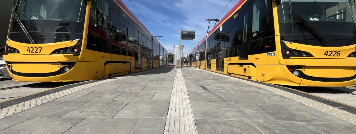 Na zdjęciu widać dwa tramwaje stojące równolegle do siebie. Pomiędzy nimi znajduje się przystanek tramwajowy.